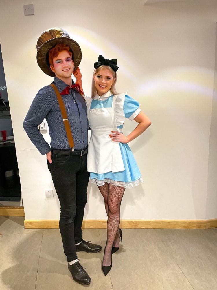 Foto de casal fantasiado de Alice e chapeleiro. Ela usa vestido azul e avental branco e ele usa chapéu marrom, camisa social e suspensório 