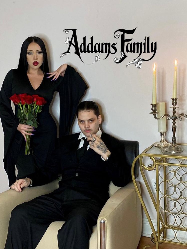 Foto de casal de Mortícia e Gomes Addams. Ela usa vestido preto e segura rosas vermelhas e ele está sentado usando terno preto 