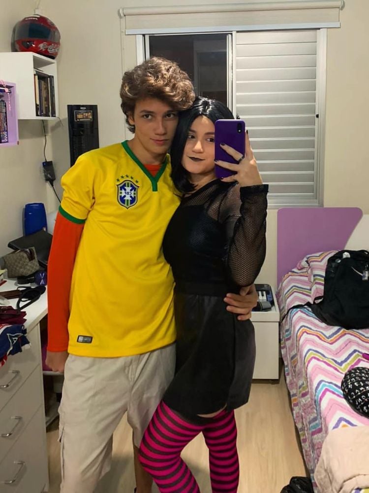 Homem com camisa amarela do Brasil, blusa laranja manga longa por baixo e bermuda bege. Mulher usando vestido preto, cabelo preto curto e maia-calça listrada 