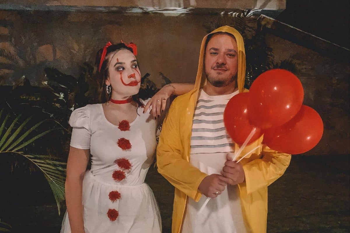 Foto de casal com fantasia de It: mulher de vestido branco com pompom vermelho e make de palhaço e homem de blusa branca, capa de chuva amarela e balões vermelhos