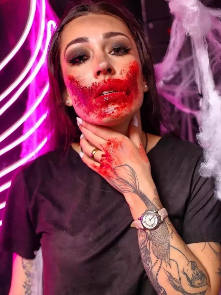 Mulher de pele clara usando blusa preta e muito sangue falso no rosto e na mão 