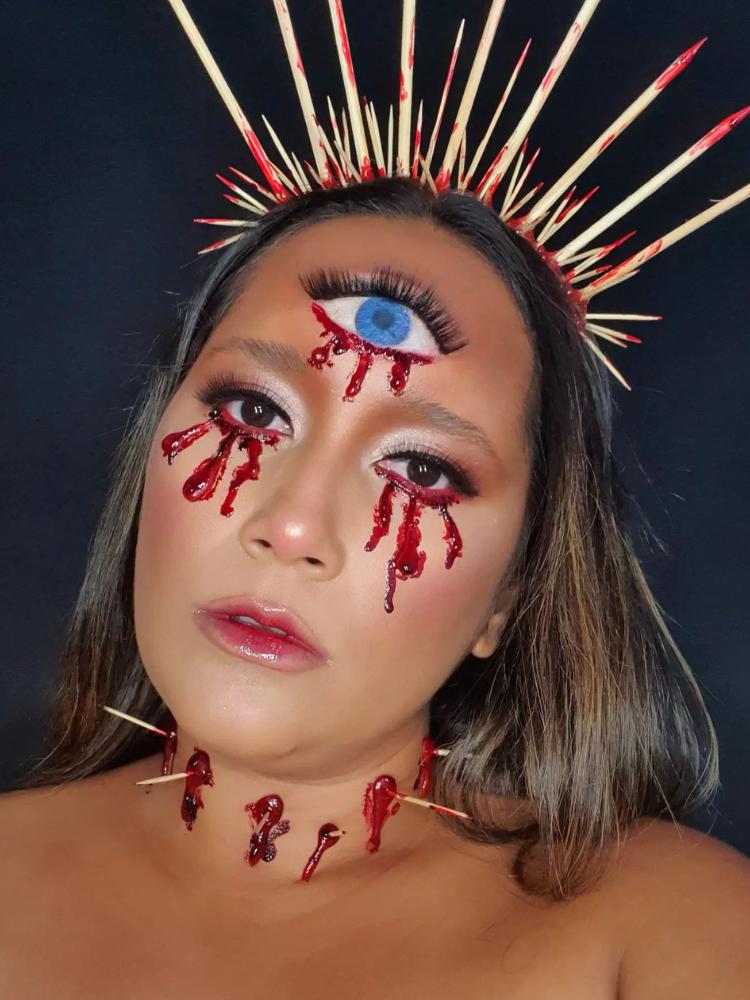 Mulher de pele clara com sangue falso escorrendo dos olhos, desenho de tolho na testa também escorrendo, palitos no pescoço simulando perfurações de estacas e tiara de palitos 