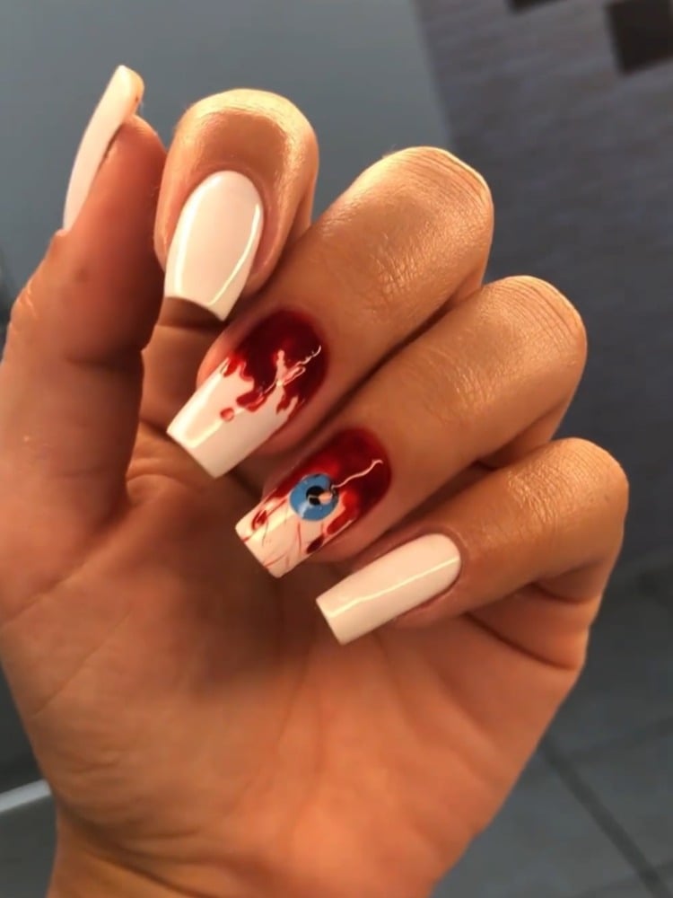 Mão de pessoa de pele clara com unhas brancas, sendo a do médio com imitação de sangue e o anelar com imitação de olho com veias e sangue