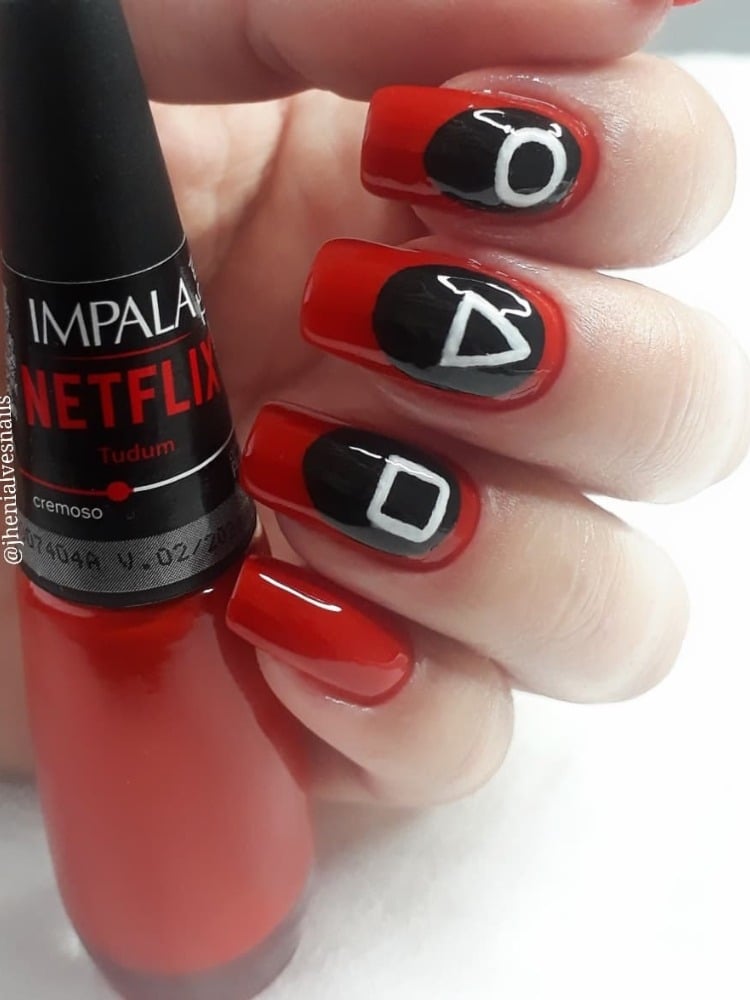 Mão de pele clara com unha média quadrada vermelha, com símbolo oval preto e quadrado, triângulo e círculo de Round 6. Além disso, há um esmalte da Impala especial Netflix, na cor vermelha