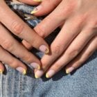 A imagem mostra mãos com unhas decoradas em estilo francesinha, exibindo pontas amarelas vibrantes e pequenos detalhes florais vermelhos. As mãos estão sobre um jeans azul claro, criando um contraste casual e moderno. A iluminação destaca a textura das unhas e a suavidade da pele.