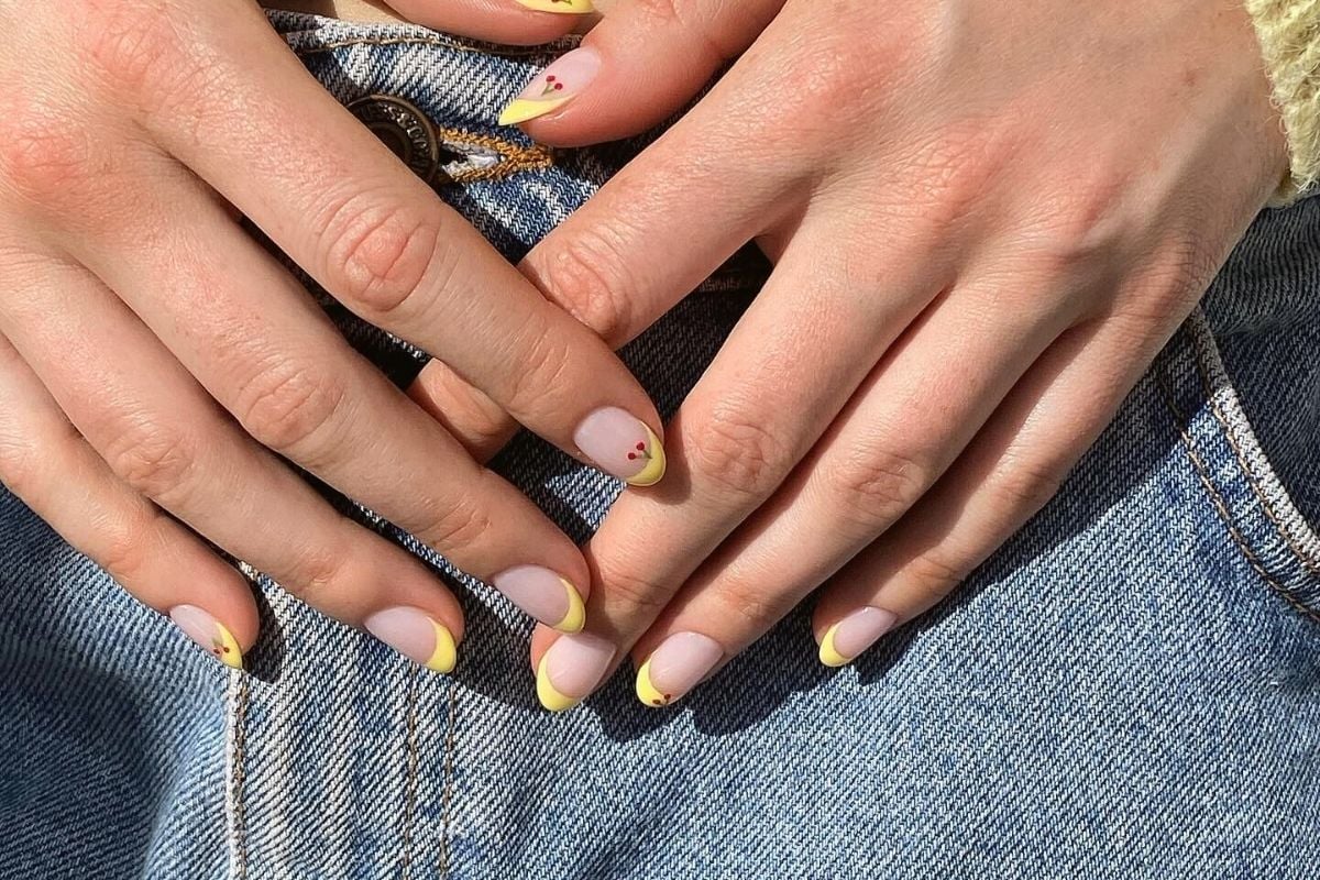 A imagem mostra mãos com unhas decoradas em estilo francesinha, exibindo pontas amarelas vibrantes e pequenos detalhes florais vermelhos. As mãos estão sobre um jeans azul claro, criando um contraste casual e moderno. A iluminação destaca a textura das unhas e a suavidade da pele.