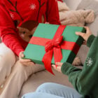 A imagem mostra duas pessoas trocando um presente embalado com papel verde e fita vermelha, simbolizando uma cena de Natal. Uma pessoa veste um suéter vermelho com tema de rena, enquanto a outra veste verde com estampa de floco de neve. Um ambiente acolhedor e festivo.