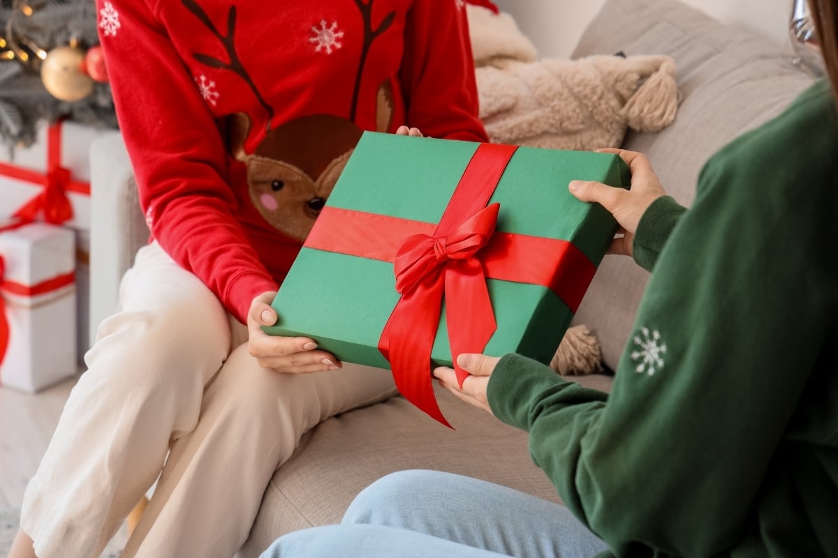 A imagem mostra duas pessoas trocando um presente embalado com papel verde e fita vermelha, simbolizando uma cena de Natal. Uma pessoa veste um suéter vermelho com tema de rena, enquanto a outra veste verde com estampa de floco de neve. Um ambiente acolhedor e festivo.