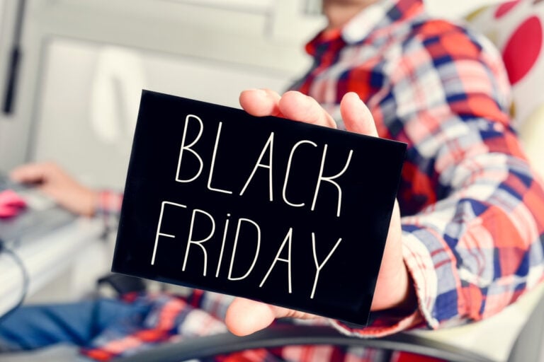 A imagem mostra uma pessoa segurando um cartão preto com as palavras "Black Friday" escritas em letras brancas. A pessoa veste uma camisa xadrez colorida e está desfocada ao fundo. O foco está no cartão, destacando a mensagem promocional típica dessa data de compras.