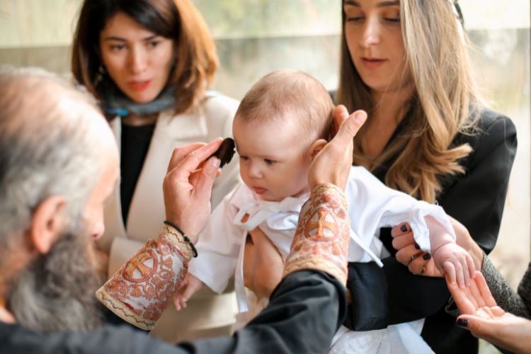 Foto de mulher de pele clara segurando bebê sendo benzido por padre e outra mulher ao fundo 