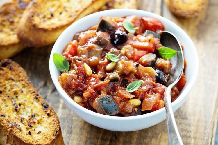 Tigela branca com caponata de berinjela e fatias de pão ao redor