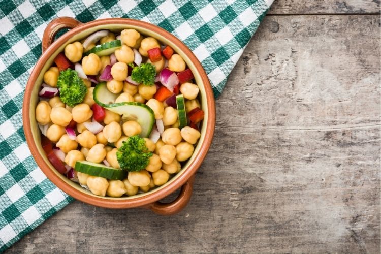 Tigela marrom com salada de grão-de-bico sobre mesa de madeira com toalha verde xadrez