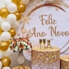 A imagem mostra uma decoração de Ano Novo com temática dourada e branca. Há balões dourados e brancos, um fundo com "Feliz Ano Novo" escrito, arranjo de flores, taças de champanhe, e mesas decoradas, criando um ambiente festivo e elegante.