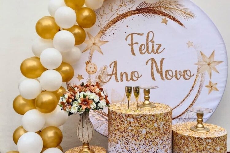 A imagem mostra uma decoração de Ano Novo com temática dourada e branca. Há balões dourados e brancos, um fundo com "Feliz Ano Novo" escrito, arranjo de flores, taças de champanhe, e mesas decoradas, criando um ambiente festivo e elegante.