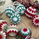 Bolas de Natal de crochê em tons vermelho, verde e branco estão sobre uma base de madeira. Decoradas com padrões florais, estão cercadas por ramos festivos, criando um cenário natalino acolhedor.