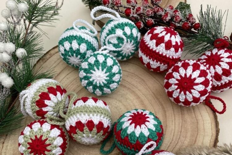 Bolas de Natal de crochê em tons vermelho, verde e branco estão sobre uma base de madeira. Decoradas com padrões florais, estão cercadas por ramos festivos, criando um cenário natalino acolhedor.