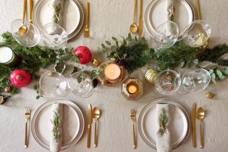 A mesa de Natal está elegantemente decorada com talheres dourados, pratos de porcelana, guardanapos com ramos de alecrim, e um assado suculento. Vegetais assados, molho de frutas vermelhas, e frutas secas completam a cena, criando uma atmosfera festiva e aconchegante.