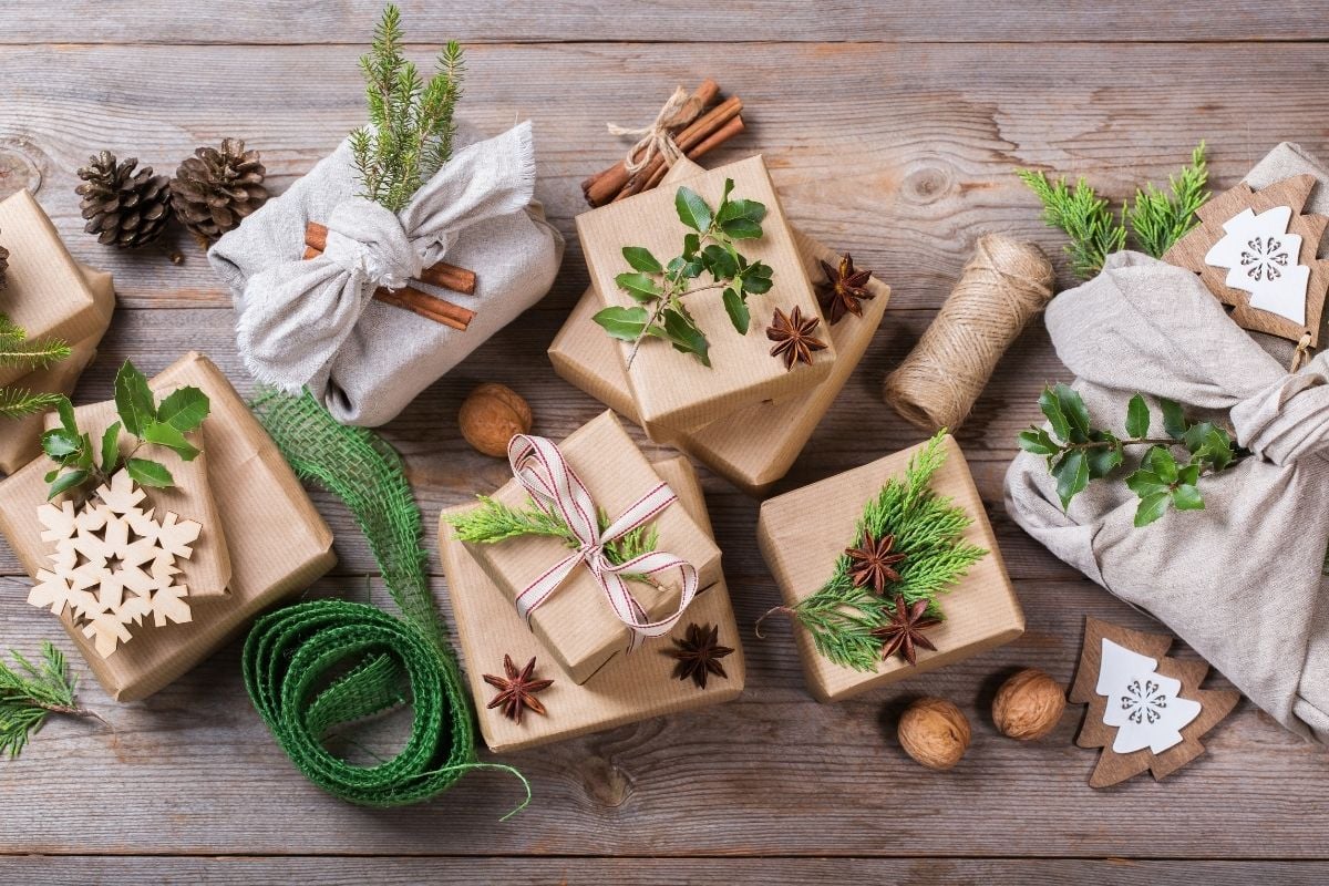 A imagem mostra presentes embrulhados em papel kraft e tecido, decorados com folhas verdes, ramos de pinheiro, canela e estrelas de anis. Há um rolo de fita verde e um barbante, criando uma estética rústica e natural sobre uma mesa de madeira.