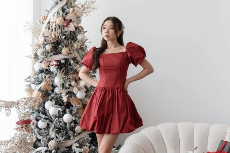 Mulher de origem asiática usando vestido vermelho com manga bufante e saia balone. Ao fundo há uma árvore de Natal