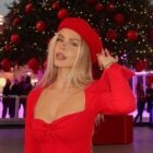 Mulher loira de pele clara usando maquiagem neutra, vestido vermelho, boina vermelha e com árvore de Natal gigante ao fundo