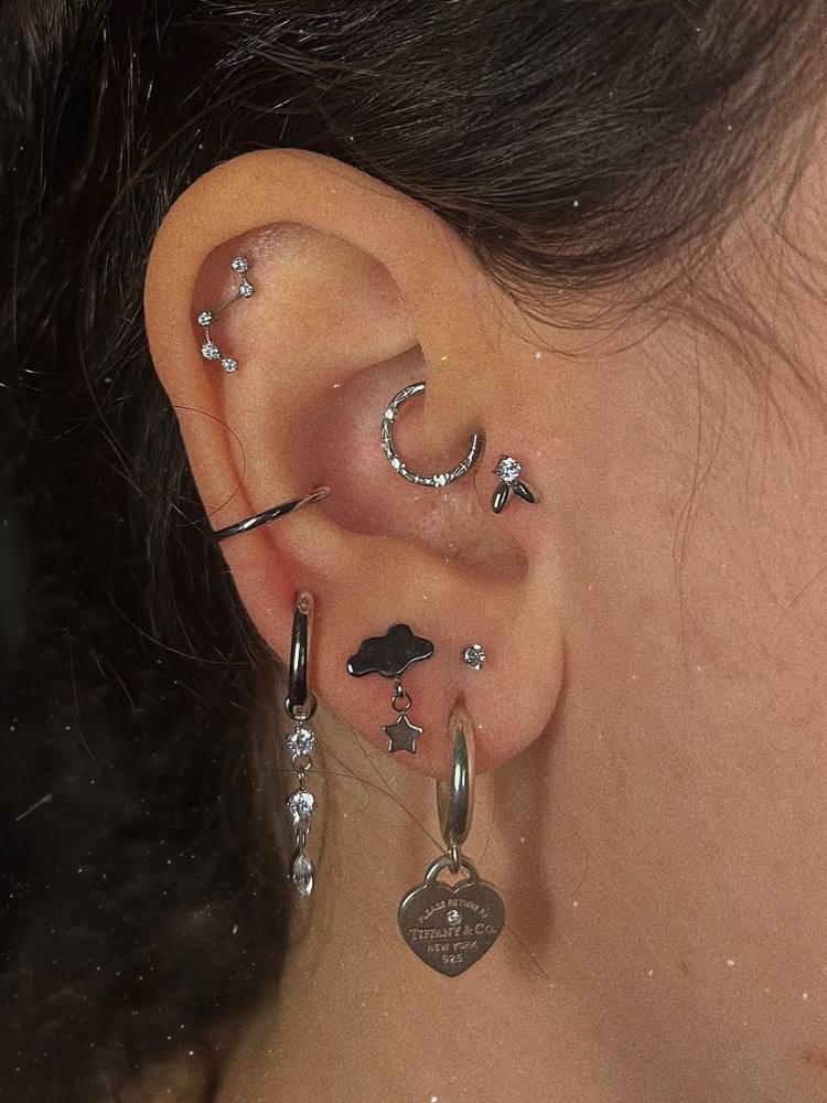 Orelha de pessoa de pele clara com constelação no flat, argola no daith, pomo de ouro no tragus, argola simples no conch, argola com pontos de luz pendurados no lóbulo, nuvem com estrela no lóbulo, argola com coração no lóbulo e ponto de luz no up lóbulo