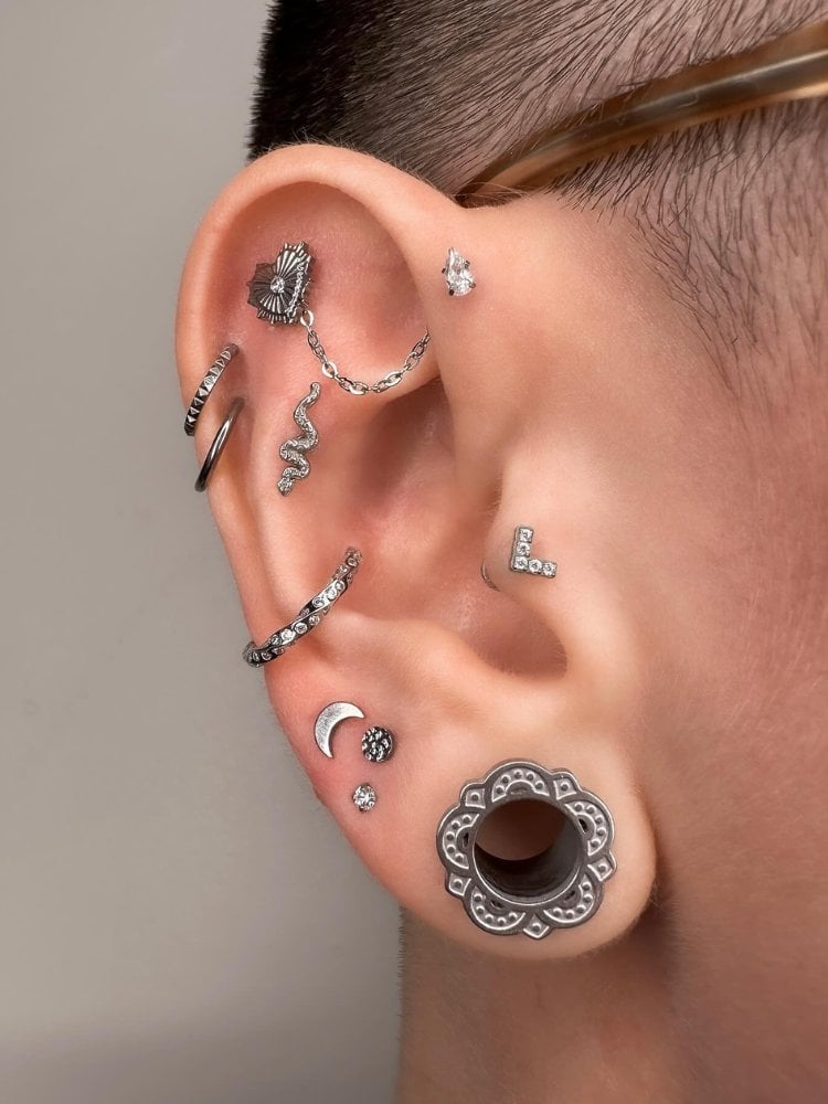 Orelha de pele clara com alargador de mandala, duplo helix com argolas, argola no conch, lua e pontos de luz abaixo do conch, V no tragus, piercing de cobra, coração na scapha com corrente até ponto de luz no anti helix