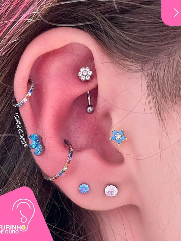 Orelha de pele clara com argola no hélix, flor no rook, jóia azul no conch, argola abaixo do conch, flor azul no tragus e pontos de luz no lóbulo