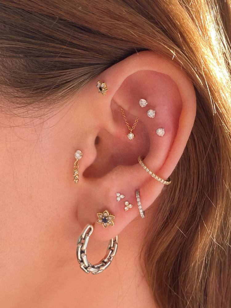 Orelha de pele clara com piercing de flor no anti helix, três pontos de luz na scapha, argola no conch, argola no primeiro lóbulo, flor no segundo, duas fores pequenas no terceiro, argola no quarto e ponto de luz com pingente no tragus 