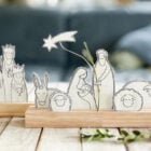 A imagem mostra um presépio minimalista com figuras de papel em bases de madeira. Os personagens incluem Maria, José, o Menino Jesus, os Três Reis Magos e animais. Uma estrela cadente destaca-se acima, enquanto ramos verdes decoram a cena, que está sobre uma superfície de madeira clara.