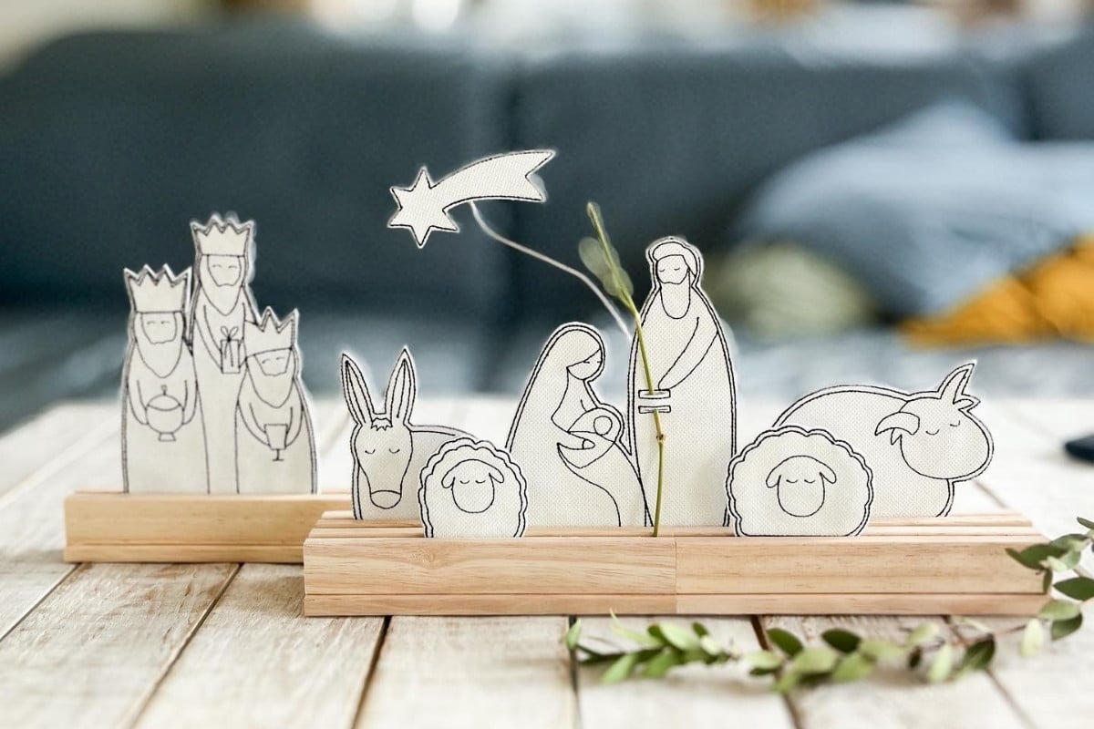 A imagem mostra um presépio minimalista com figuras de papel em bases de madeira. Os personagens incluem Maria, José, o Menino Jesus, os Três Reis Magos e animais. Uma estrela cadente destaca-se acima, enquanto ramos verdes decoram a cena, que está sobre uma superfície de madeira clara.