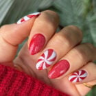 A imagem mostra uma mão com unhas decoradas para o Natal. As unhas têm um design alternado de glitter vermelho e listras brancas em espiral, semelhante a doces de hortelã. A pessoa veste um suéter vermelho, criando uma estética festiva.