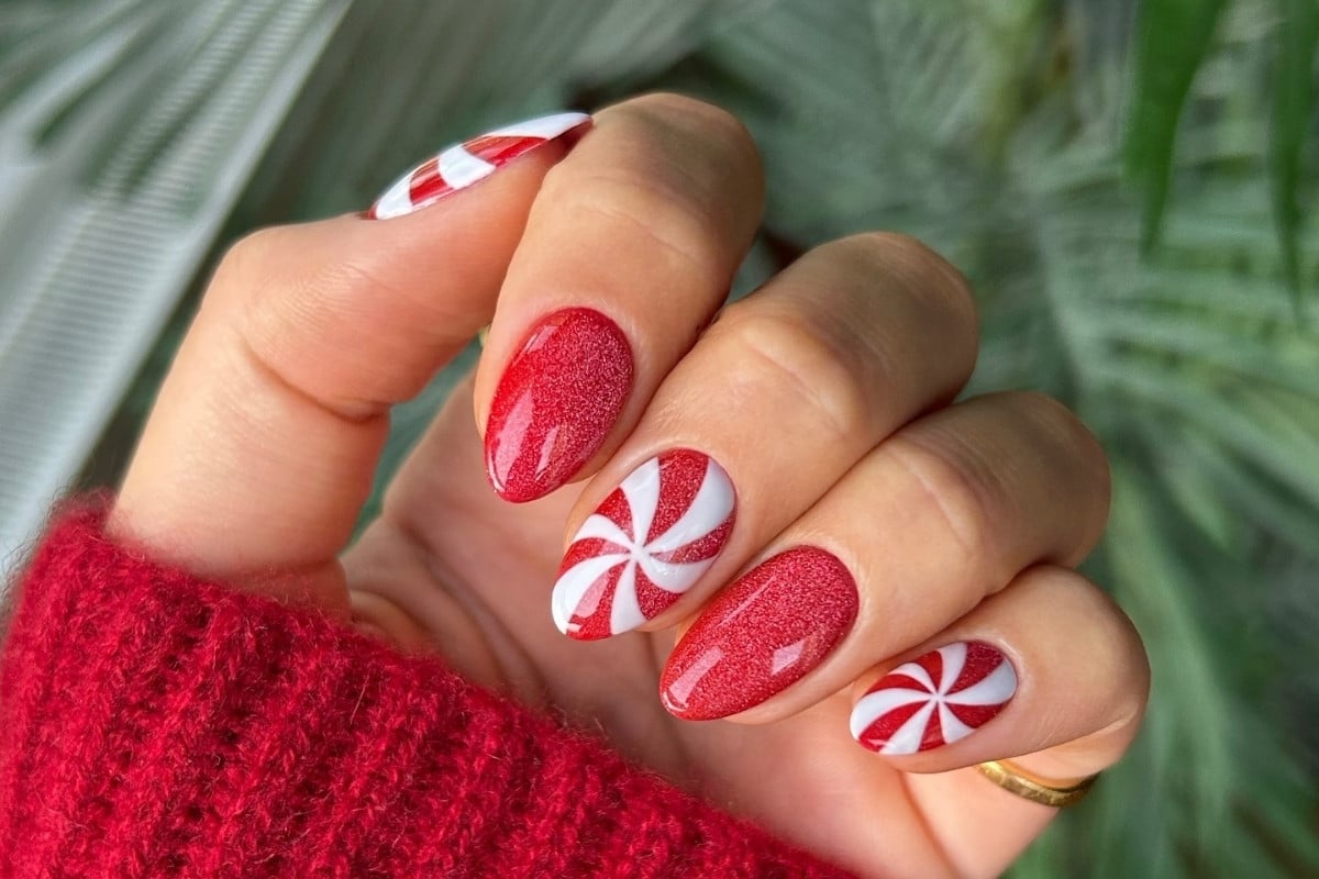 A imagem mostra uma mão com unhas decoradas para o Natal. As unhas têm um design alternado de glitter vermelho e listras brancas em espiral, semelhante a doces de hortelã. A pessoa veste um suéter vermelho, criando uma estética festiva.