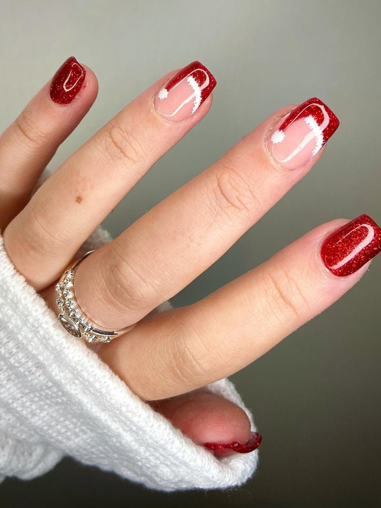 Mão de pele clara com unha média quadrada com unhas vermelhas e outras bege com nail art de gorro de papai noel