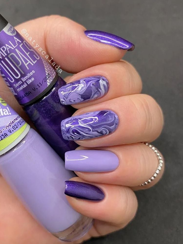 Mão de pele clara segurando esmaltes roxos e unhas grandes quadradas com marmorizado roxo 