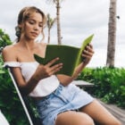 A mulher está sentada, lendo um livro verde. Ela veste uma blusa branca e shorts jeans, em um ambiente ao ar livre com vegetação ao fundo. O clima parece tranquilo, com céu nublado e palmeiras ao redor.