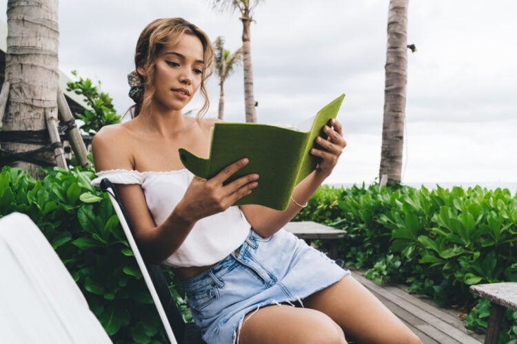 A mulher está sentada, lendo um livro verde. Ela veste uma blusa branca e shorts jeans, em um ambiente ao ar livre com vegetação ao fundo. O clima parece tranquilo, com céu nublado e palmeiras ao redor.