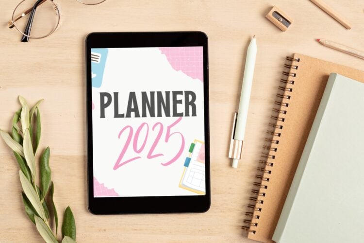 Mesa bege com cadernos, planta, óculos, caneta e tablet com planner 2025 em rosa, azul e preto