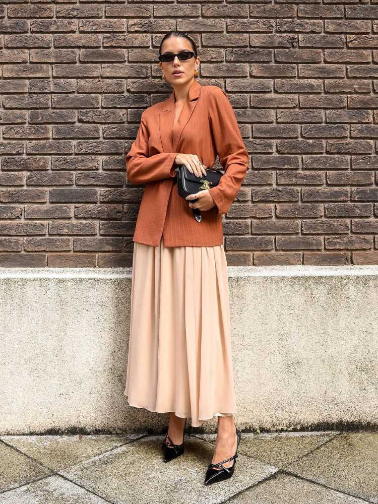 Mulher de pele clara usando blazer marrom e saia bege, em frente à parede de tijolos à vista 