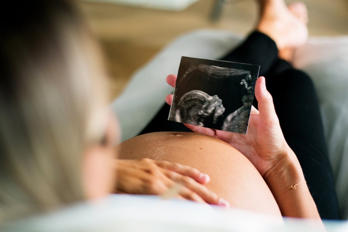 Mulher de pele clara deitada com a mão na barriga grávida, calça preta e segurando foto de ultrassom