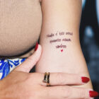 A imagem apresenta uma tatuagem delicada no braço de uma mulher com a frase: "Nada é tão nosso quanto nossos sonhos". Abaixo da frase, há um pequeno coração vermelho. Detalhes incluem unhas pintadas e acessórios elegantes.