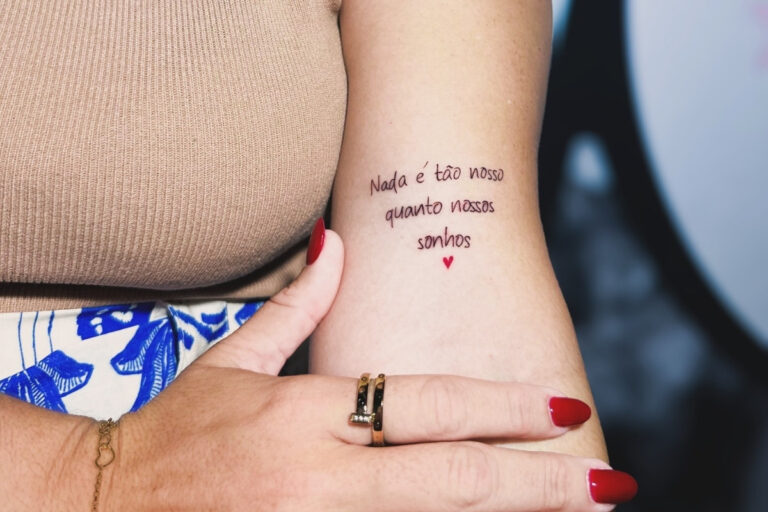 A imagem apresenta uma tatuagem delicada no braço de uma mulher com a frase: "Nada é tão nosso quanto nossos sonhos". Abaixo da frase, há um pequeno coração vermelho. Detalhes incluem unhas pintadas e acessórios elegantes.