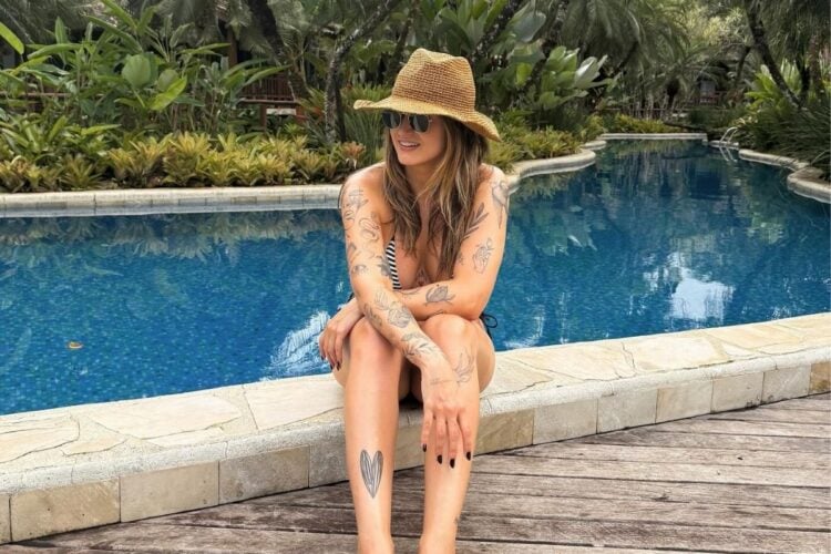 Mari Palma sentada à beira da piscina com suas tatuagens dos braços e pernas expostas