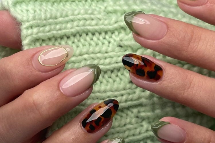 Uma mão com unhas artísticas exibe diferentes designs: pontas verdes, um padrão de tartaruga e um contorno dourado. O fundo apresenta um tecido verde tricotado. As unhas têm um acabamento brilhante e elegante, criando um visual moderno e sofisticado.