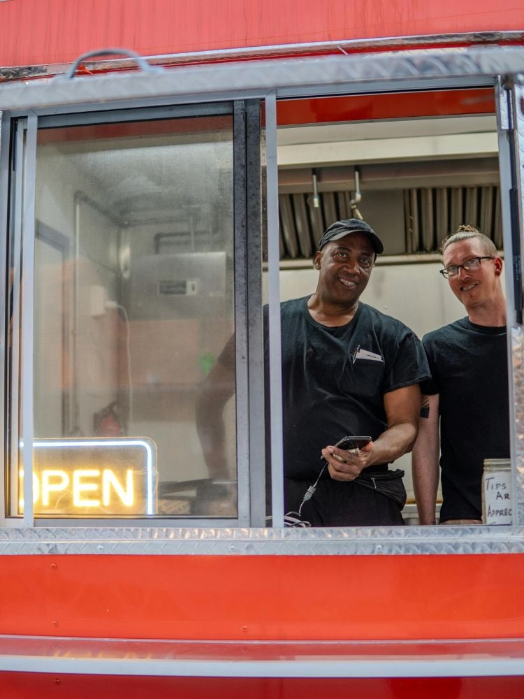 Foto de dois trabalhadores de rede de fast food na janela do drive through. Os dois sorriem e usam uniformes pretos
