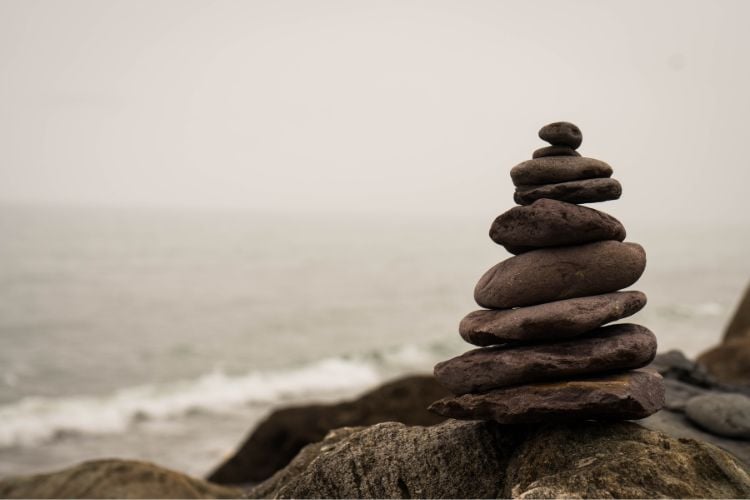 Foto de pilha de pedras em frente ao mar em dia nublado, representando equilíbrio entre a espiritualidade e as demandas da vida cotidiana
