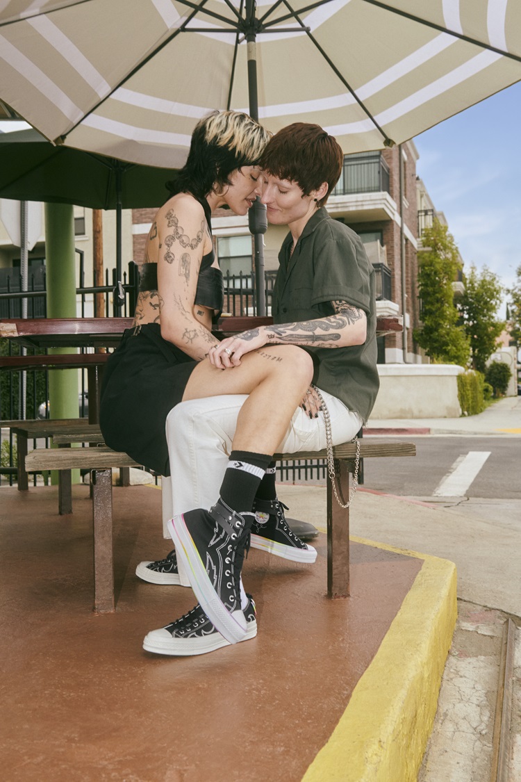 Foto de casal sentado frente a frente em banco de madeira. As duas mulheres usam modelos de tênis da coleção Converse Pride 2024