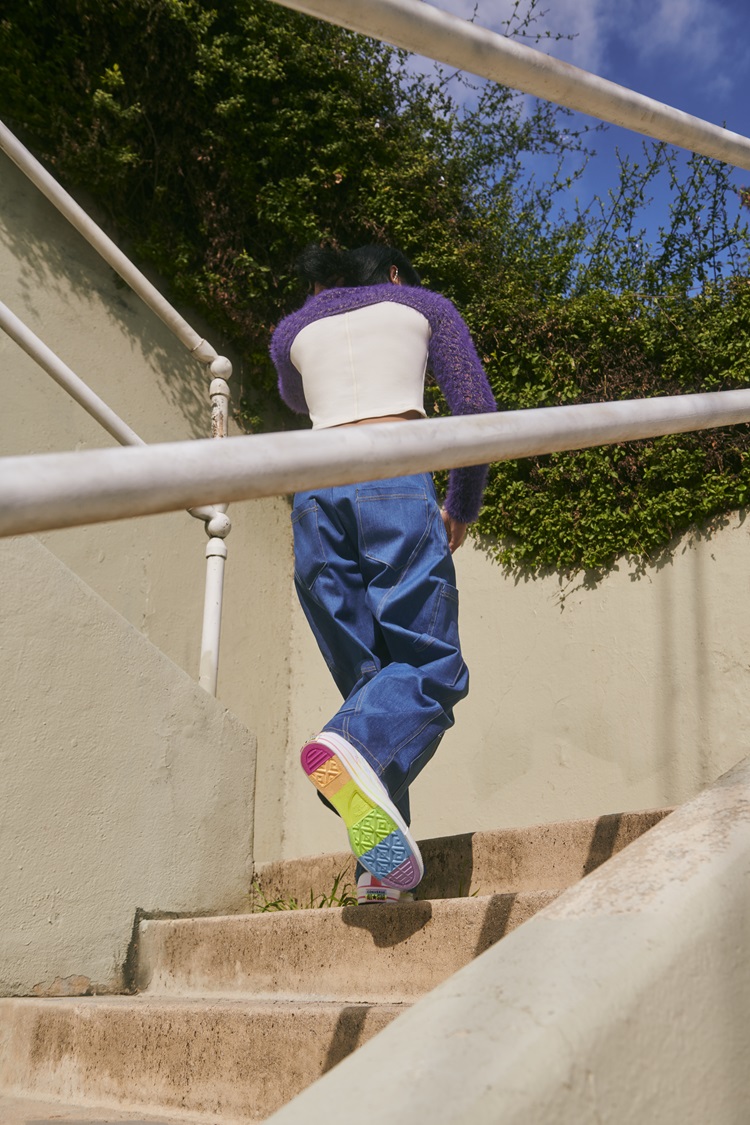 Foto de pessoa de costas subindo escada usando bolero de lã, cropped branco, calça jeans e tênis da coleção Converse Pride 2024