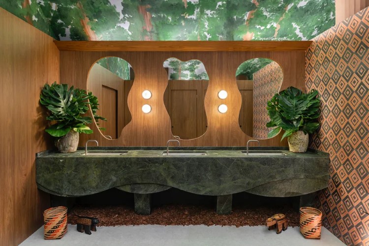 A imagem mostra um banheiro com decoração moderna e elementos naturais. Há uma pia de mármore verde com três torneiras, espelhos ovais, plantas, iluminação embutida e papel de parede geométrico.