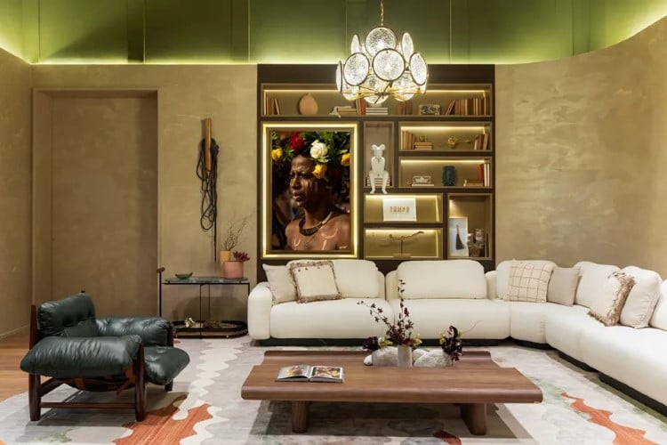 A imagem exibe uma sala de estar elegante com sofá branco, poltrona verde, mesa de centro de madeira, estante embutida com objetos decorativos e iluminação de lustre.