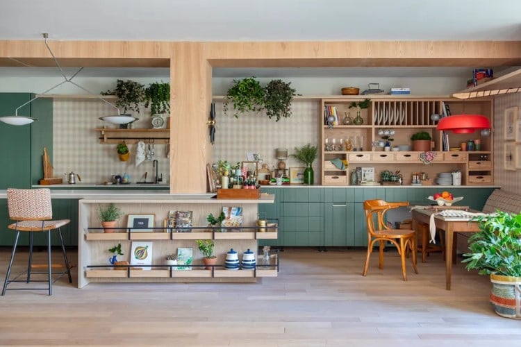 A cozinha moderna exibe prateleiras de madeira, armários verdes e plantas. Há uma área de jantar com cadeiras, uma mesa posta para dois e itens decorativos pelo espaço.